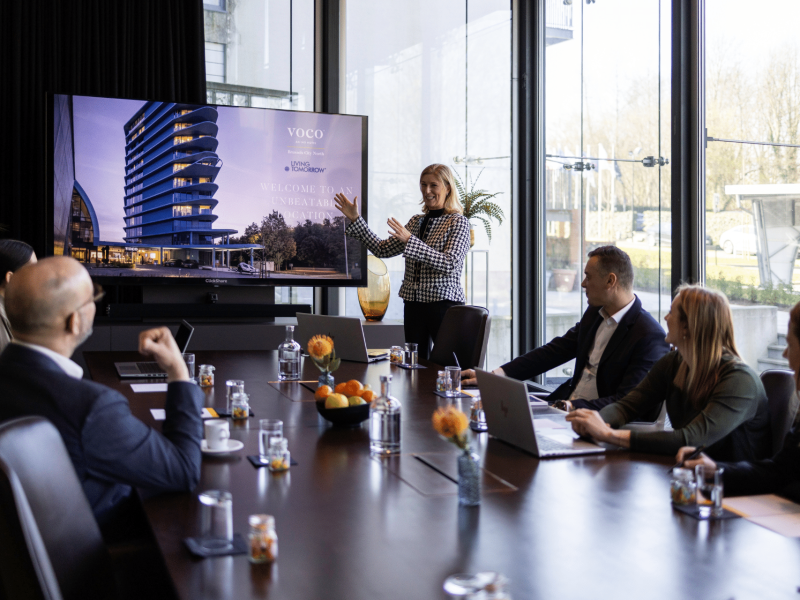 woman smiling and presenting next to the TV screen to a group of people sitting around the table